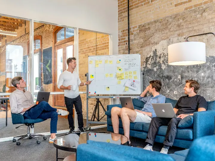 three men sitting while using laptops and watching man beside whiteboard